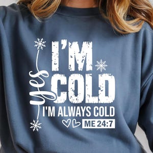 May include: A blue sweatshirt with the text "I'M COLD I'M ALWAYS COLD ME 24:7" in white. The sweatshirt has a crew neck and long sleeves. The text is decorated with snowflakes and the word "yes".