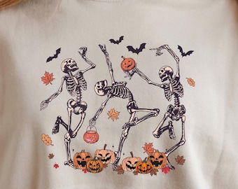 May include: Beige sweatshirt featuring a Halloween-themed graphic. The design includes dancing skeletons, pumpkins, bats, and autumn leaves. The illustration is in shades of black, orange, and brown. The sweatshirt is a casual, long-sleeved garment.