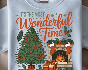 May include: A light blue sweatshirt with a festive Christmas design. The graphic includes a Christmas tree, fireplace, and the text "It's the Most Wonderful Time" in orange and green. The sweatshirt is a Gildan brand.