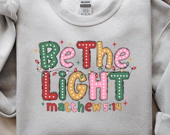 May include: A light gray crewneck sweatshirt featuring the text Be The Light Matthew 5:14 in colorful, polka-dotted bubble letters. The design is accented with a string of small, multi-colored Christmas lights and tiny red star shapes.
