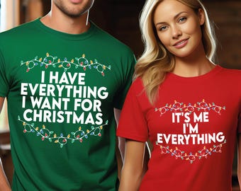 May include: A man and woman wearing matching Christmas-themed t-shirts. The green shirt says "I HAVE EVERYTHING I WANT FOR CHRISTMAS" and the red shirt says "IT'S ME I'M EVERYTHING". Both shirts have a string of Christmas lights design.