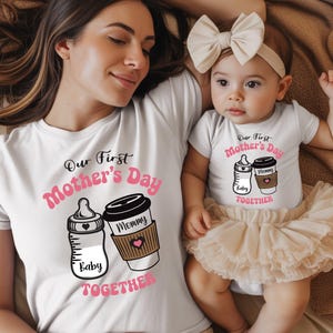 May include: White t-shirts with matching graphics for a mother and baby. The shirts feature a baby bottle and coffee cup design with the text "Our First Mother's Day Baby Mommy Together". The baby wears a tutu.