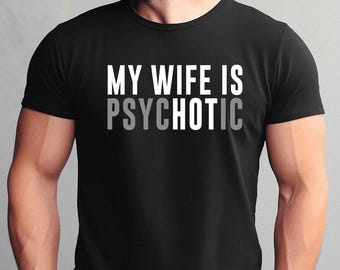 May include: A black short-sleeved t-shirt featuring the white and grey text MY WIFE IS PSYCHOTIC printed on the chest. The word PSYCHOTIC is displayed with the letters PSYCH and TIC in grey, while the letter O is in white.