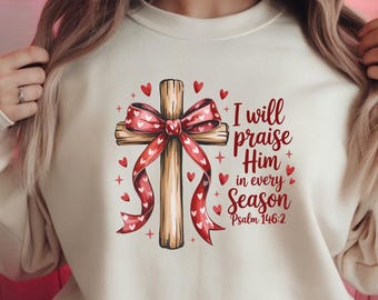 May include: A cream-colored crewneck sweatshirt featuring a graphic of a wooden cross adorned with a red heart-patterned ribbon. The text reads, I will praise Him in every Season Psalm 146:2, surrounded by small red hearts and star accents.