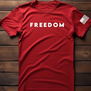 May include: A red t-shirt with the word "FREEDOM" printed in white across the chest. The shirt has a small American flag on the left sleeve. The shirt is displayed on a dark wood background.