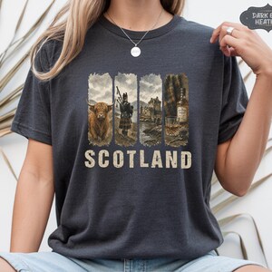 May include: Dark grey heather t-shirt with a Scotland graphic. The graphic features a Highland cow, a bagpiper, a castle, and a bottle of whiskey with a glass. The word "SCOTLAND" is printed in a distressed font below the images.