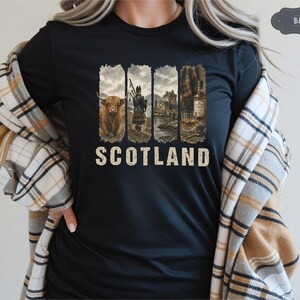 May include: Black t-shirt with a graphic design featuring Scottish imagery. The design includes a Highland cow, a bagpiper, a castle, and a whiskey bottle with a glass. The word "SCOTLAND" is printed in a vintage font.