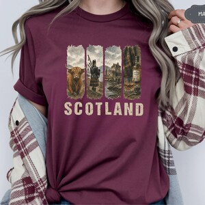 May include: Maroon t-shirt featuring a vintage-style design with four vertical panels. The panels depict a Highland cow, a bagpiper, a castle, and a whiskey bottle with a glass. The word "SCOTLAND" is printed in a distressed font.