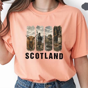 May include: Peach-colored t-shirt featuring a graphic design with the word "SCOTLAND" in bold black letters. The design includes images of a Highland cow, a bagpiper, a castle, and a whiskey bottle with a glass. The shirt is made of a soft material.