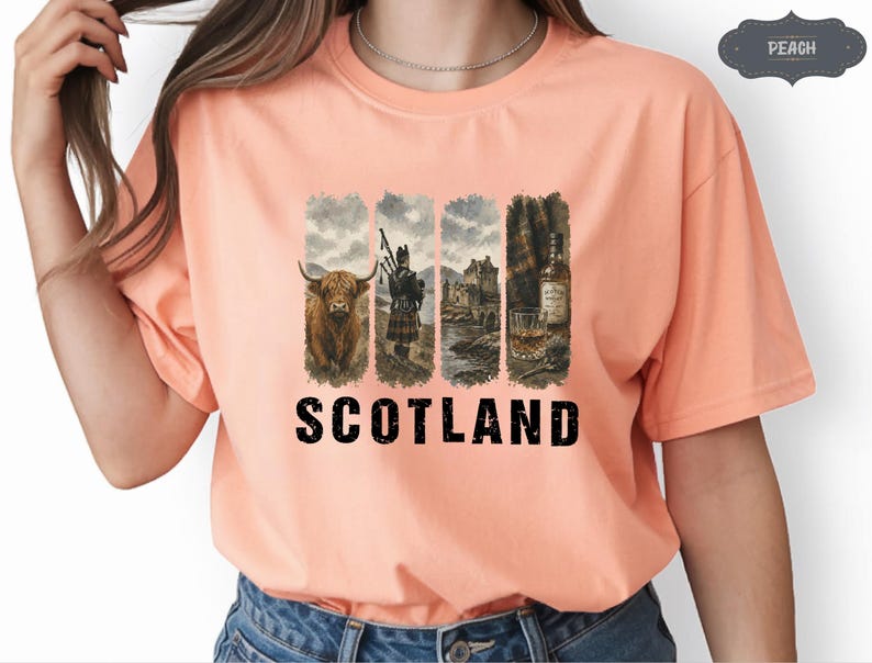 May include: Peach-colored t-shirt featuring a graphic design with the word "SCOTLAND" in bold black letters. The design includes images of a Highland cow, a bagpiper, a castle, and a whiskey bottle with a glass. The shirt is made of a soft material.