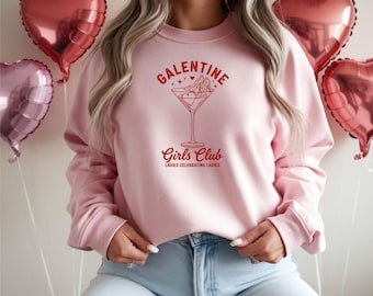 May include: A light pink sweatshirt with the text "GALENTINE Girls Club Ladies Celebrating Ladies" in red. The design features a martini glass with high heels inside. Heart-shaped balloons are in the background.