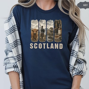 May include: Navy blue t-shirt featuring a distressed graphic with images of a Highland cow, a bagpiper, a castle, and a whiskey bottle. The word "SCOTLAND" is printed in a vintage font below the images. The shirt is a solid navy color.