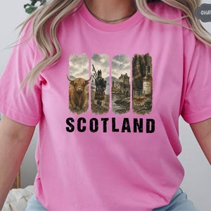 May include: Pink t-shirt featuring a graphic design with four vertical panels. The panels depict a Highland cow, a bagpiper, a castle, and a whiskey bottle and glass. The word "SCOTLAND" is printed in bold black letters.