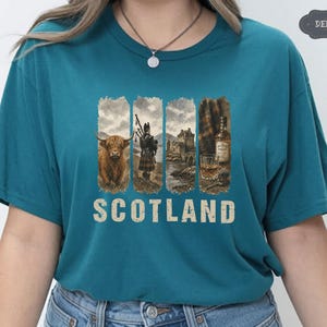 May include: Teal t-shirt featuring a graphic design with Scottish imagery. The design includes a Highland cow, a bagpiper, a castle, and a glass of whiskey. The word "SCOTLAND" is printed in a distressed font below the images.