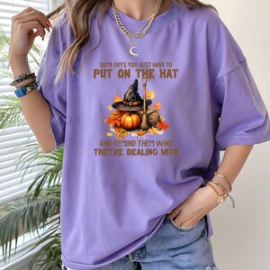 May include: A lilac-colored t-shirt with the text "SOME DAYS YOU JUST HAVE TO PUT ON THE HAT AND REMIND THEM WHO THEY'RE DEALING WITH." The graphic features a witch's hat, pumpkin, broom, and autumn leaves.