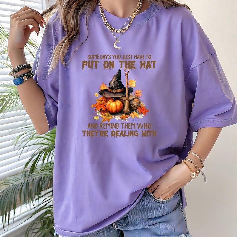 May include: A lilac-colored t-shirt with the text "SOME DAYS YOU JUST HAVE TO PUT ON THE HAT AND REMIND THEM WHO THEY'RE DEALING WITH." The graphic features a witch's hat, pumpkin, broom, and autumn leaves.