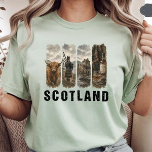 May include: A light green t-shirt featuring a graphic design with Scottish imagery. The design includes a Highland cow, a bagpiper, a castle, and a glass of whiskey. The word "SCOTLAND" is printed in bold, black letters.