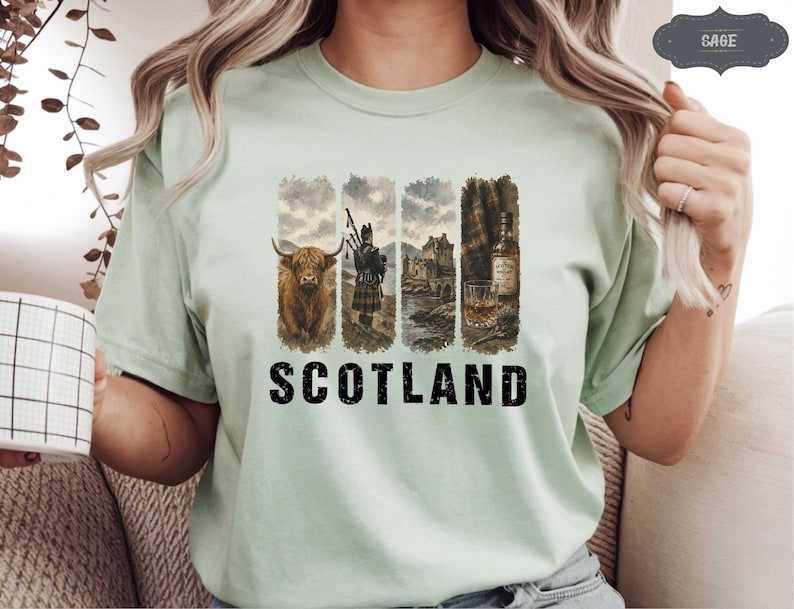 May include: A light green t-shirt featuring a graphic design with Scottish imagery. The design includes a Highland cow, a bagpiper, a castle, and a glass of whiskey. The word "SCOTLAND" is printed in bold, black letters.