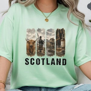 May include: A light green t-shirt with a graphic design featuring a Highland cow, a bagpiper, a castle, and a whiskey bottle. The word "SCOTLAND" is printed in bold, black letters below the images.