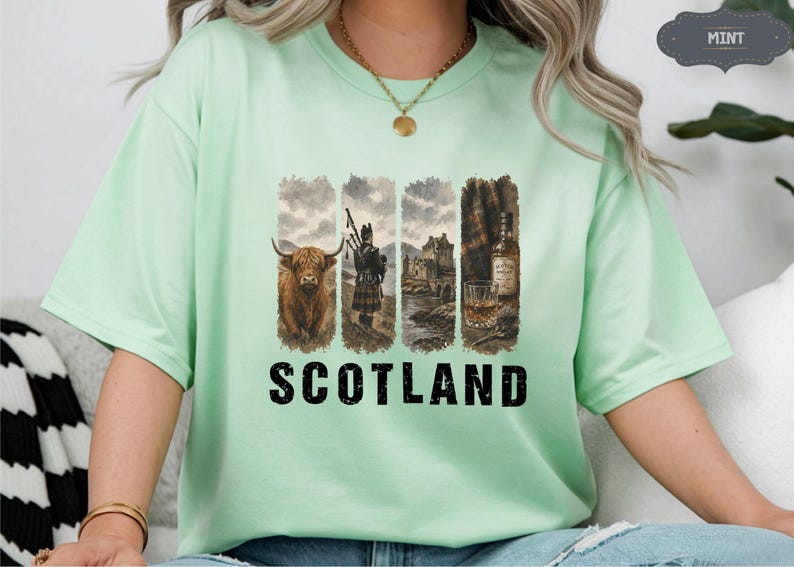 May include: A light green t-shirt with a graphic design featuring a Highland cow, a bagpiper, a castle, and a whiskey bottle. The word "SCOTLAND" is printed in bold, black letters below the images.