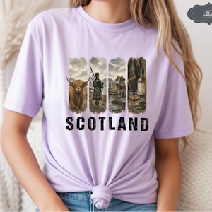 May include: Lilac t-shirt with a graphic featuring Scottish imagery. The design includes a Highland cow, a bagpiper, a castle, and a whiskey bottle with a glass. The word "SCOTLAND" is printed in bold, black letters below the images.