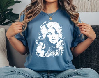 May include: A blue jean colored t-shirt featuring a white graphic print of a woman with voluminous hair and hoop earrings. The shirt has a crew neckline and short sleeves. A small label in the top right corner displays the text BLUE JEAN.