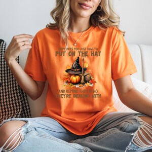 May include: Orange t-shirt with a Halloween graphic. The graphic features a witch's hat, pumpkin, and broom with the text "SOME DAYS YOU JUST HAVE TO PUT ON THE HAT AND REMIND THEM WHO THEY'RE DEALING WITH."