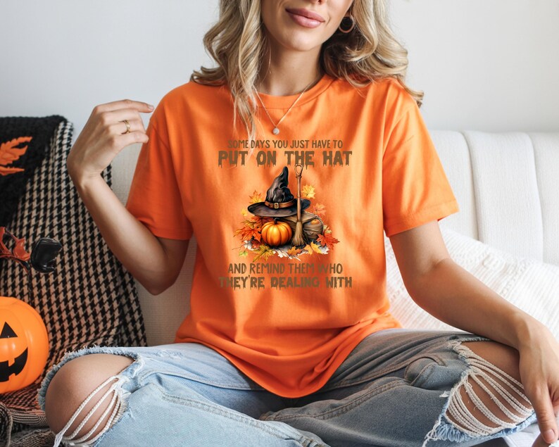 May include: Orange t-shirt with a Halloween graphic. The graphic features a witch's hat, pumpkin, and broom with the text "SOME DAYS YOU JUST HAVE TO PUT ON THE HAT AND REMIND THEM WHO THEY'RE DEALING WITH."