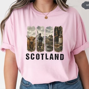 May include: A light pink t-shirt featuring a graphic of Scotland. The graphic includes a Highland cow, a bagpiper, a castle, and a bottle of whiskey. The word "SCOTLAND" is printed in bold black letters below the graphic.