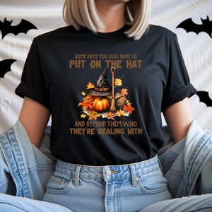 May include: Black t-shirt featuring a Halloween-themed graphic. The design includes a witch's hat, pumpkin, broom, and autumn leaves. Text on the shirt reads, "SOME DAYS YOU JUST HAVE TO PUT ON THE HAT AND REMIND THEM WHO THEY'RE DEALING WITH."