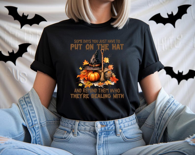 May include: Black t-shirt featuring a Halloween-themed graphic. The design includes a witch's hat, pumpkin, broom, and autumn leaves. Text on the shirt reads, "SOME DAYS YOU JUST HAVE TO PUT ON THE HAT AND REMIND THEM WHO THEY'RE DEALING WITH."