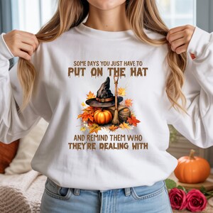 May include: White sweatshirt with a graphic featuring a witch's hat, broom, and pumpkin surrounded by autumn leaves. The text reads, "SOME DAYS YOU JUST HAVE TO PUT ON THE HAT AND REMIND THEM WHO THEY'RE DEALING WITH."