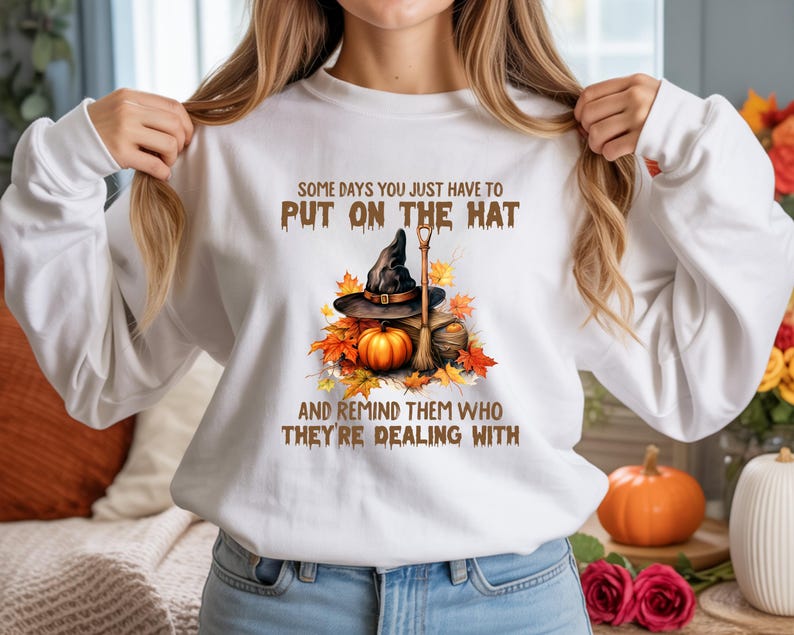 May include: White sweatshirt with a graphic featuring a witch's hat, broom, and pumpkin surrounded by autumn leaves. The text reads, "SOME DAYS YOU JUST HAVE TO PUT ON THE HAT AND REMIND THEM WHO THEY'RE DEALING WITH."