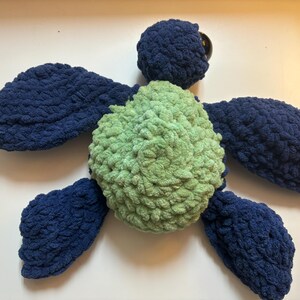 May include: A handmade plush turtle in shades of blue and green. The turtle's body is a soft, textured green, with a dark blue head, flippers, and tail. The eyes are dark brown. A cute, cuddly, and whimsical toy.