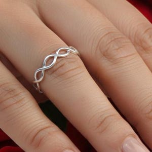 May include: A silver ring with an infinity symbol design is displayed on a hand with manicured nails. The ring is set against a backdrop of vibrant red roses, creating a romantic and elegant aesthetic.