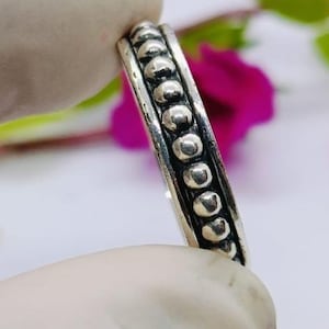 Puede incluir: Un anillo de plata con un diseño de cuentas. El anillo se sostiene entre dos dedos enguantados, con un fondo borroso de pétalos de flores coloridos y hojas verdes. La banda del anillo está adornada con pequeñas cuentas espaciadas.