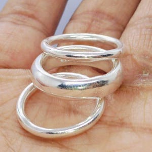 Op de afbeelding: Drie zilveren ringen van verschillende breedtes zijn op een hand gestapeld. De ringen zijn glad en glanzend en reflecteren licht. De ringen zijn waarschijnlijk gemaakt van sterling zilver of een vergelijkbaar metaal. De ringen hebben een eenvoudig, klassiek ontwerp.