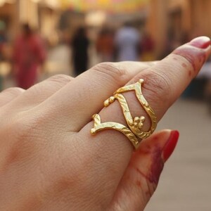 May include: A gold-colored ring with an open design, featuring a floral accent. The ring is worn on a finger, with a blurred background of a building and people. The ring's design is intricate and detailed.