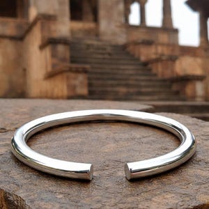 May include: A shiny, silver-colored open cuff bracelet. The bracelet is a thick, rounded band with a smooth, polished surface. The ends of the bracelet are open, creating a gap. The bracelet is resting on a textured, brown stone surface.