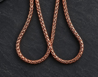 May include: A rose gold-toned wheat chain necklace featuring a tightly woven, braided metal link design. The chain has a polished, reflective surface and is arranged in two symmetrical loops, showcasing the intricate, repeating pattern of the links.