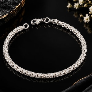 May include: A silver-toned wheat chain bracelet featuring a polished, braided metal link design. The bracelet is constructed with a secure lobster claw clasp and a circular jump ring at the ends, showcasing a continuous, intricate woven pattern.