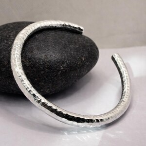 May include: A silver cuff bracelet with a hammered texture. The open-ended bracelet is displayed on a black stone against a white background. The bracelet's design features a textured surface, reflecting light.