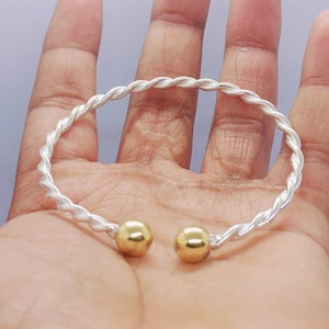 May include: A silver-toned, twisted-rope style bracelet with gold-colored ball ends. The bracelet is a bangle style, designed to slip over the hand. The metal has a polished finish, reflecting light. The bracelet is a piece of jewelry.