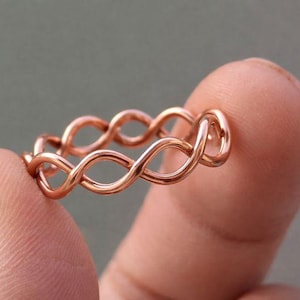 May include: A copper-colored ring with a continuous, interwoven design. The ring's pattern features a series of connected loops, creating a flowing, openwork aesthetic. The jewelry is held between two fingers, showcasing its delicate construction.