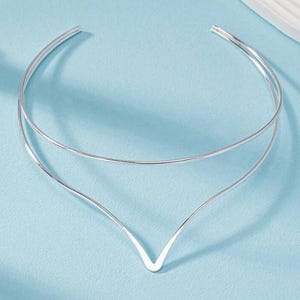 May include: A silver-colored choker necklace with a double-layered design. The top layer is a simple, curved band, while the lower layer forms a V-shape at the front. The necklace is displayed against a light blue background.