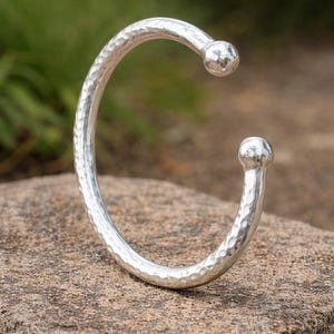 May include: A silver cuff bracelet with a hammered finish. The open design has rounded ball ends. The bracelet is shown on a textured stone surface, with a blurred green backdrop.