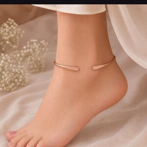 May include: A rose gold-toned metal anklet featuring a minimalist open-ended design with slightly flattened, tapered ends. The band has a smooth, polished finish and curves around the ankle, creating a simple and clean aesthetic for the jewelry piece.