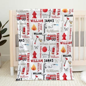 May include: A white baby blanket with a repeating pattern of red fire trucks, fire hydrants, flames, and the name "William James" in various fonts. The blanket is draped over a crib.