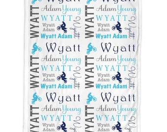 May include: A white fleece blanket featuring a repeating pattern of the name Wyatt, the name Adam Young, and a graphic of a person riding a dirt bike. The text and graphics are printed in shades of dark blue, light blue, and grey across the fabric.