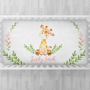 May include: A white crib sheet with a watercolor illustration of a giraffe sitting on a bed of green leaves and pink flowers. The sheet is personalized with the name "Emily Sarah" in pink lettering.